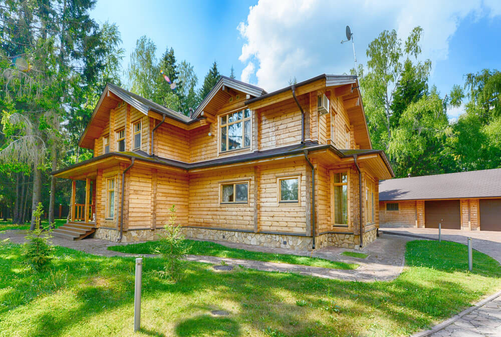 Une maison en bois de plain-pied avec un toit sombre et des fenêtres blanches, entourée d'herbe haute et avec une forêt de conifères en arrière-plan sous un ciel bleu clair.